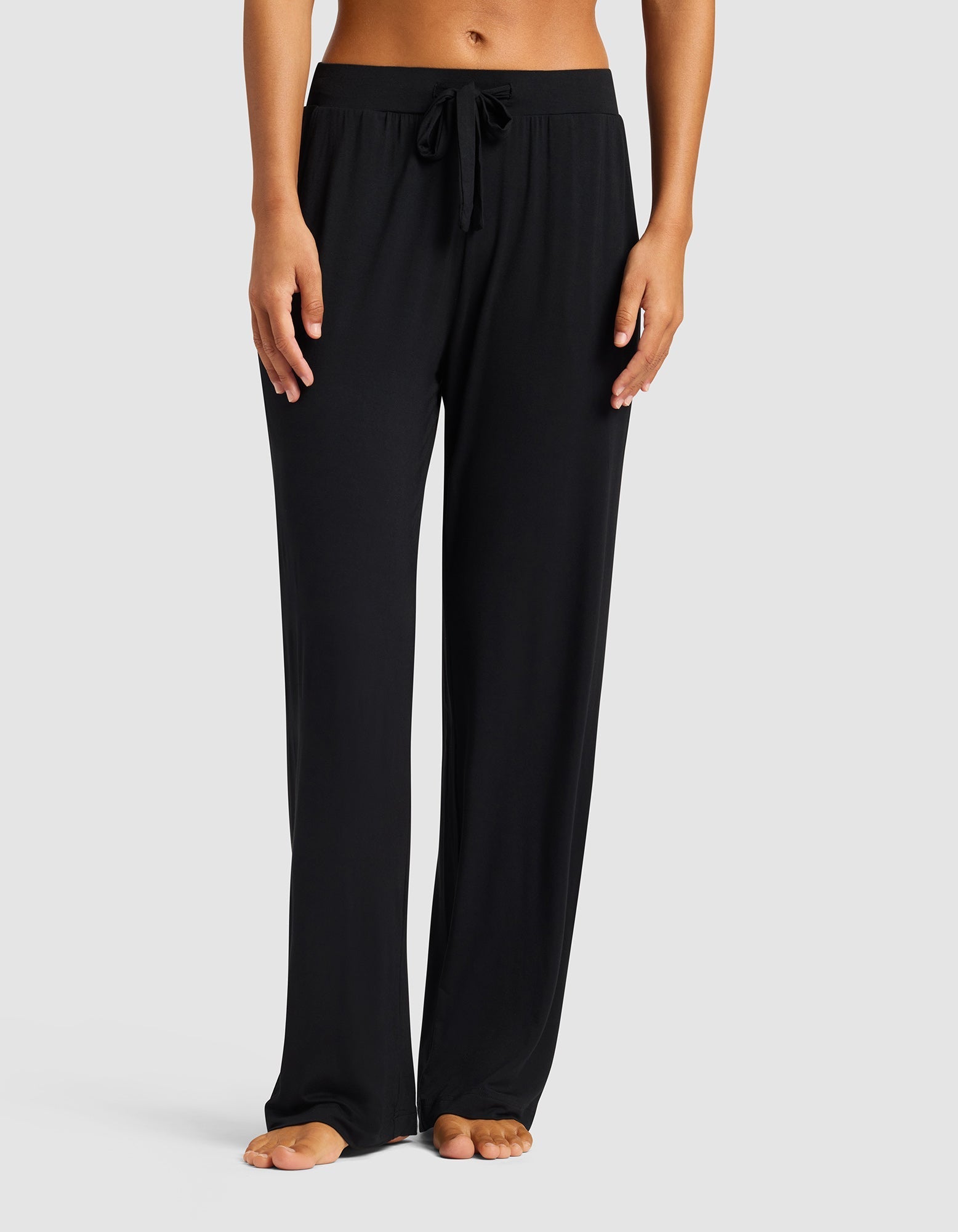 A woman stands barefoot against a plain light gray background, wearing Cozy Earth’s Women's Bamboo Stretch-Knit Pant in black with a drawstring waist and relaxed, comfortable fit.|Color:Black