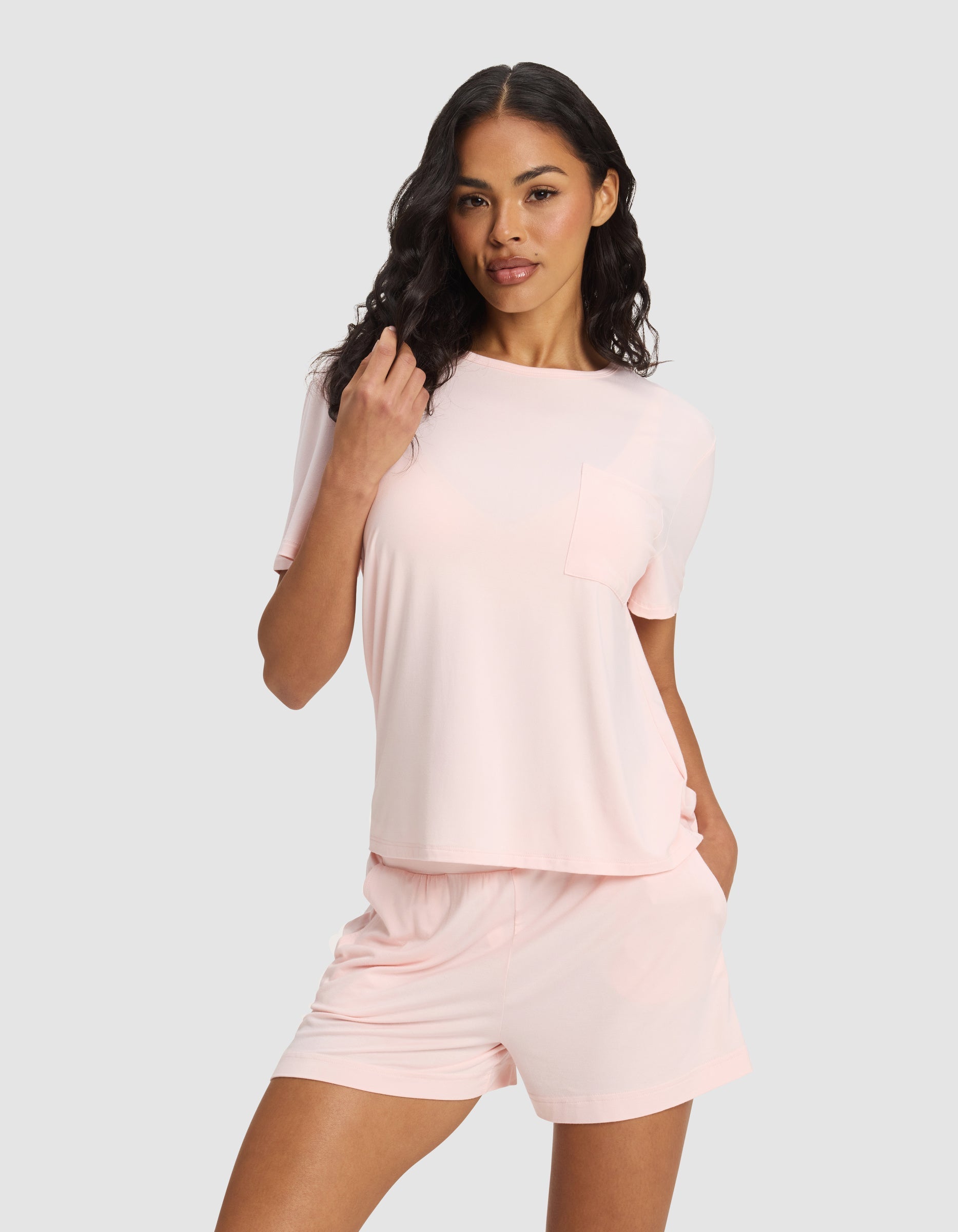 A woman with long, wavy dark hair and a relaxed, confident look stands against a plain background wearing Cozy Earth's Women's Bamboo Stretch-Knit Lounge Tee in light pink with a pocket, paired with matching pink shorts.|Color:Blossom
